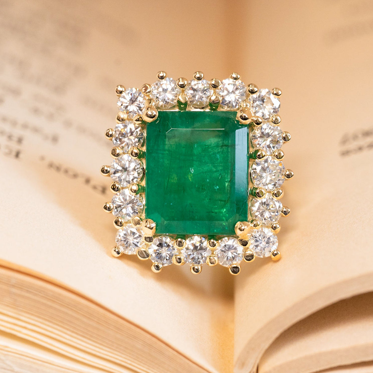 Emerald and diamond cluster cocktail ring featuring a vibrant emerald-cut emerald surrounded by brilliant-cut diamonds in yellow gold.