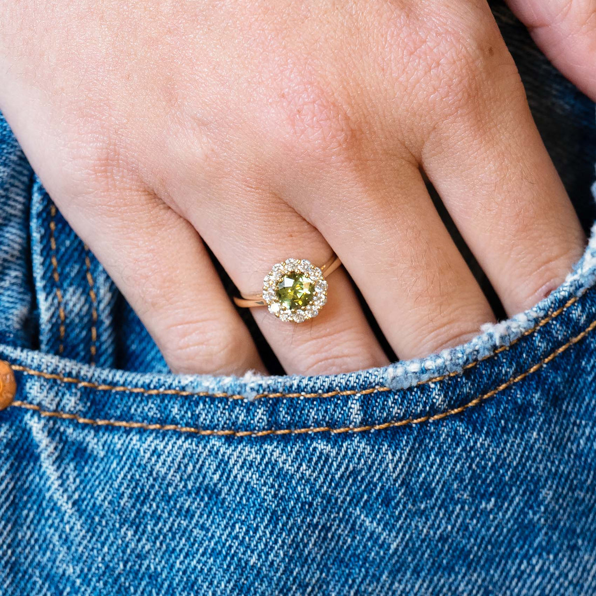 Green sapphire and diamond cluster engagement ring worn with hand resting in denim pocket.