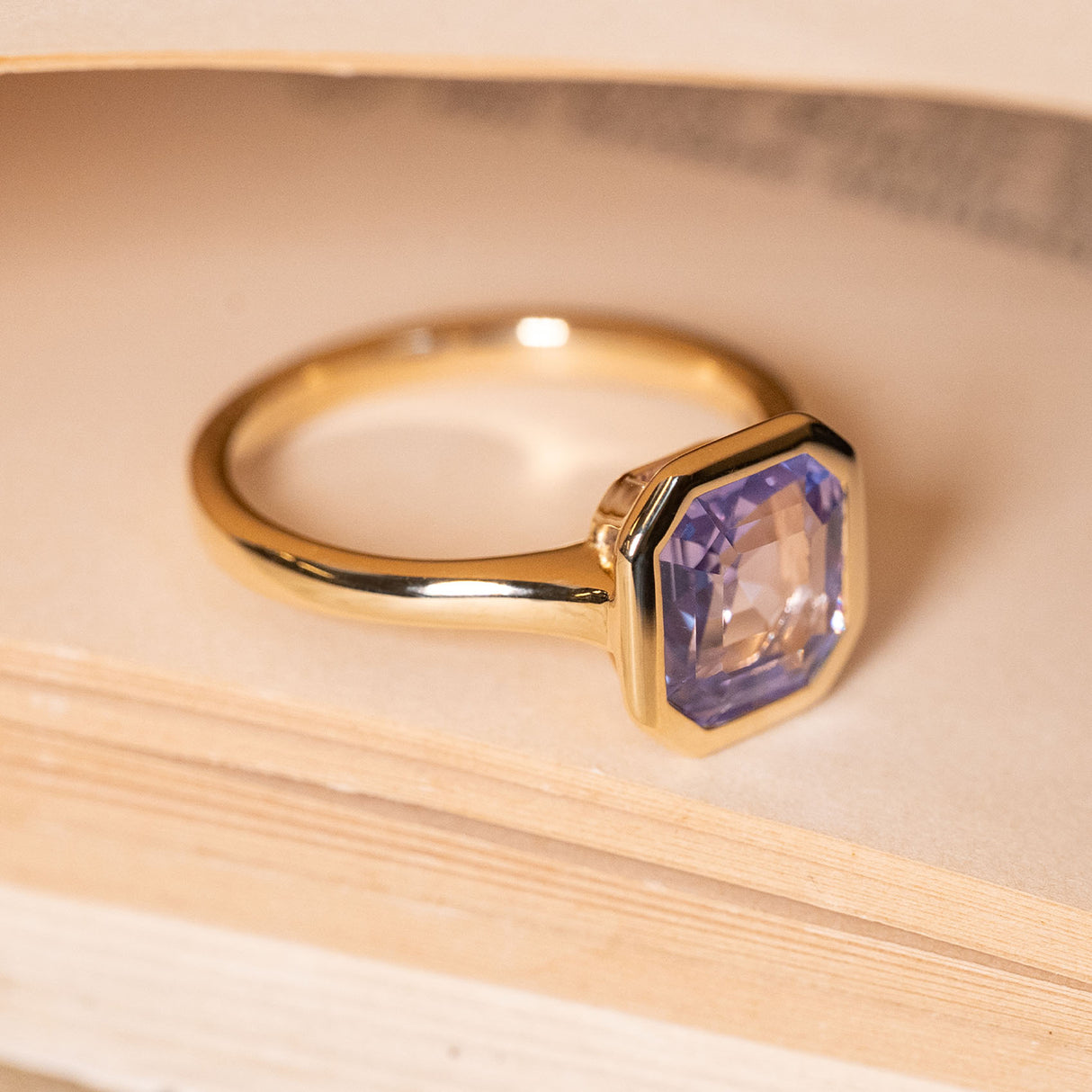 Antique-inspired lavender sapphire solitaire ring showcasing an emerald-cut lavender sapphire in a yellow gold rub-over setting.