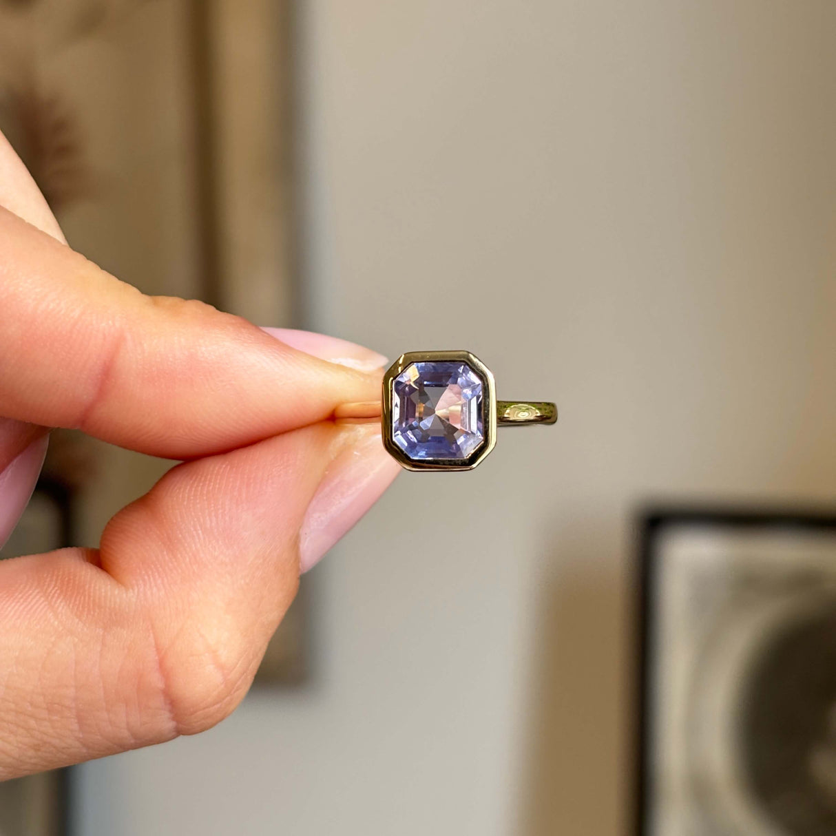 Antique-inspired square-cut lavender sapphire in a smooth yellow gold rub-over setting, held between fingers against a softly blurred vintage interior background.