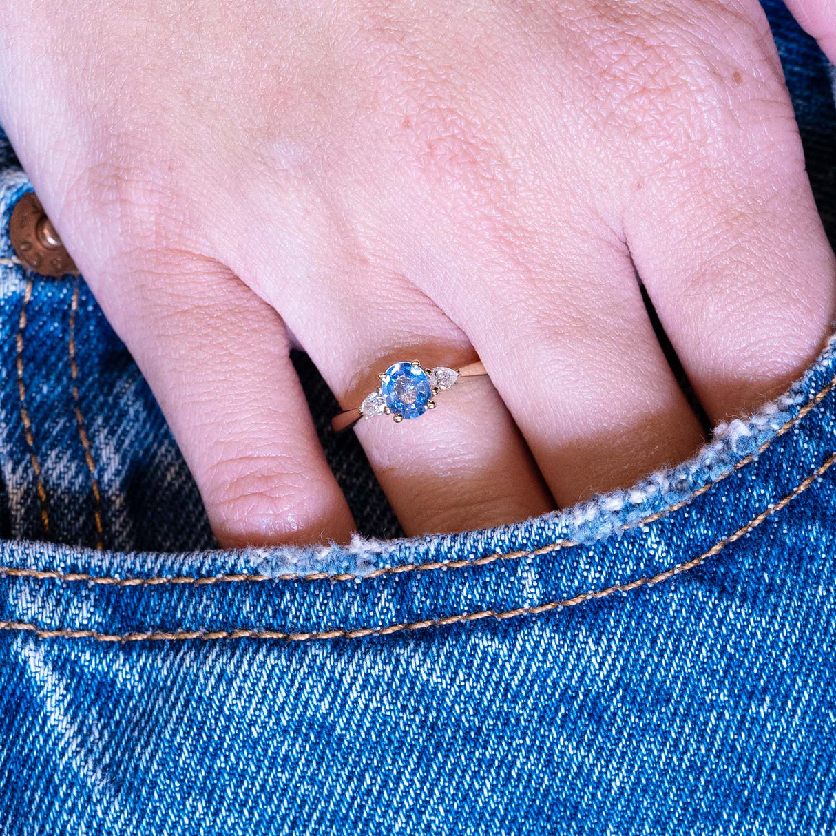 Light blue sapphire and diamond three-stone engagement ring worn on hand with denim pocket styling.