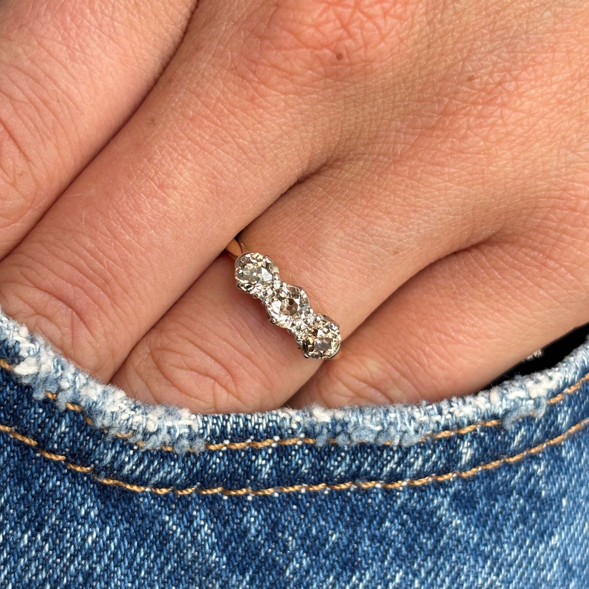 Antique Edwardian three-stone diamond ring in yellow gold styled on hand against blue denim, showcasing old-cut brown, pink and champagne coloured diamonds with subtle antique brilliance.
