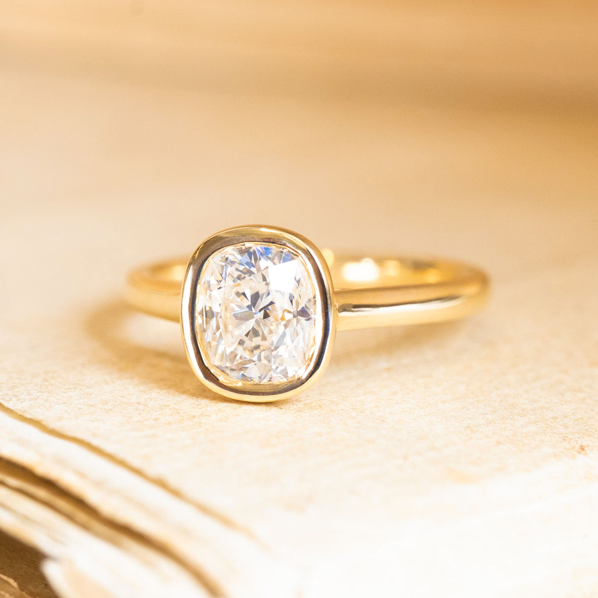 Cushion old-cut diamond solitaire engagement ring with a bezel-set diamond and polished yellow gold band.