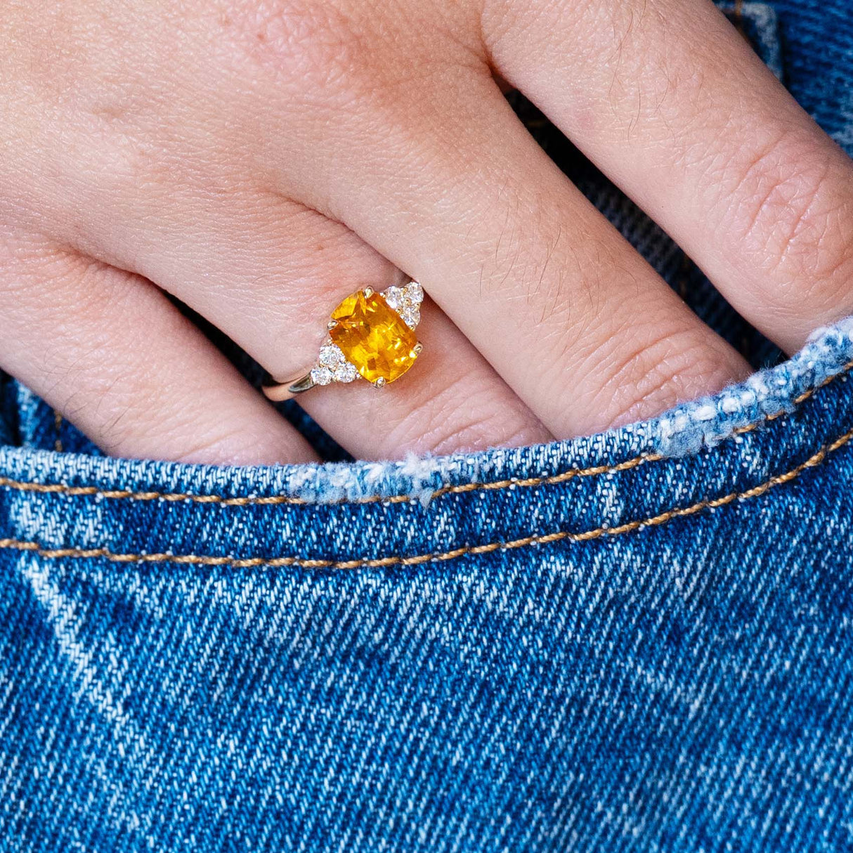Close-up of a hand wearing a ring with an orange sapphire, with a blue denim pocket detail.