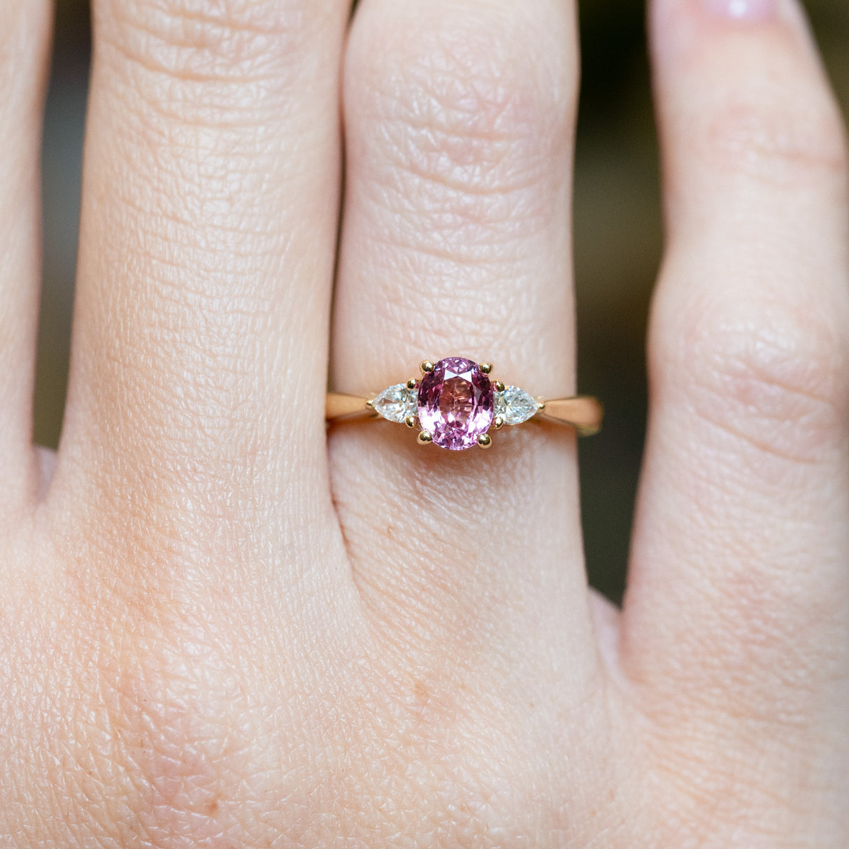 Pink sapphire and diamond three-stone engagement ring worn on finger with pear-shaped diamond side stones.