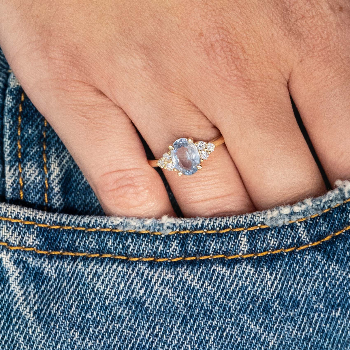 Pale blue sapphire and diamond cluster tre-foil engagement ring worn on hand resting in denim pocket.