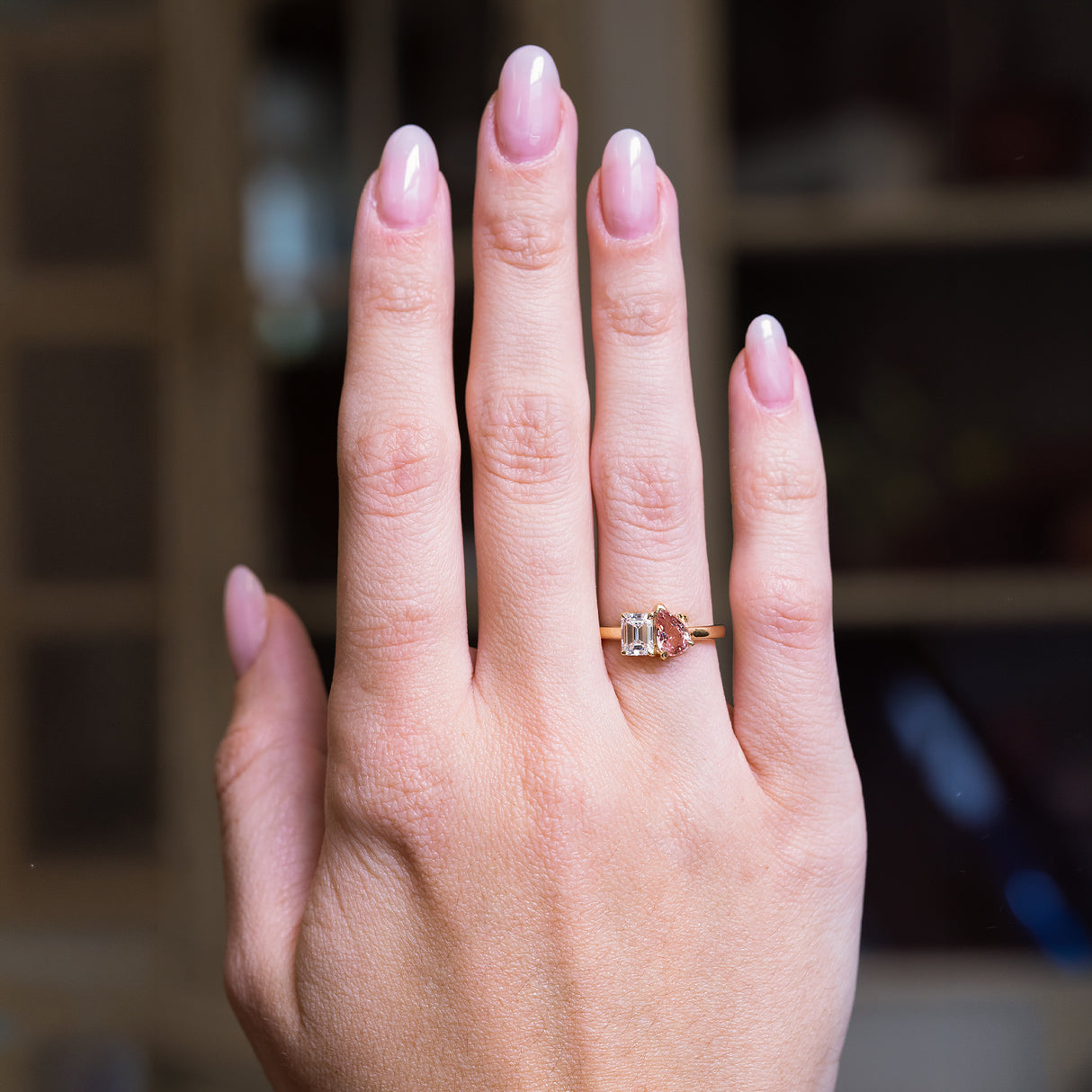 Peach coloured sapphire and diamond Toi et Moi engagement ring worn on hand with gold band.