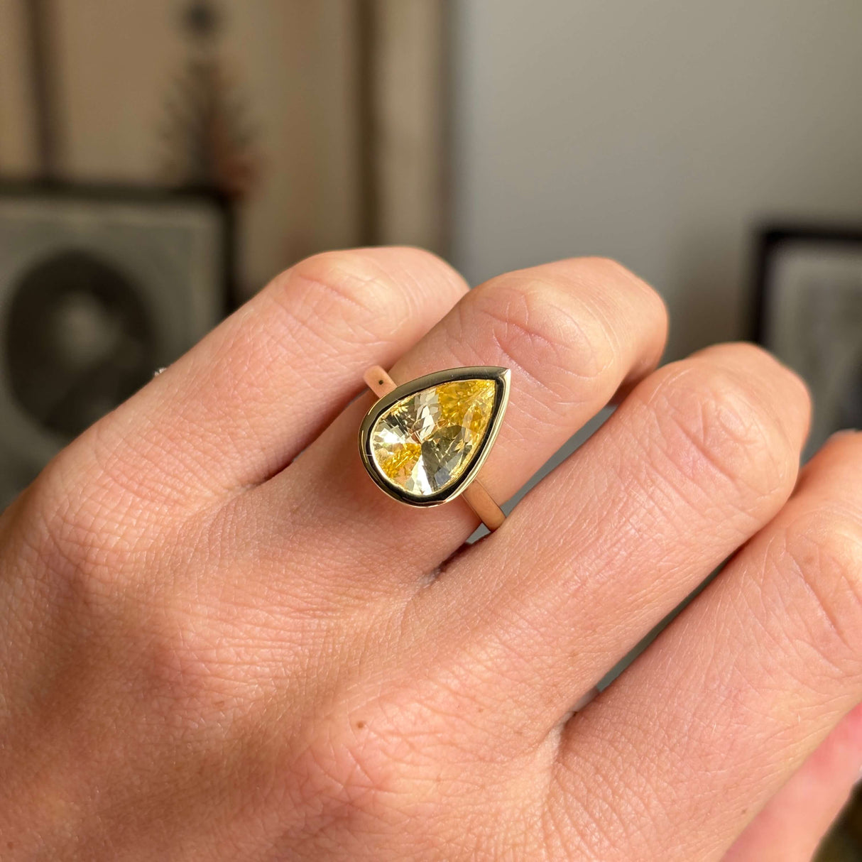Close-up of an antique-inspired yellow gold sapphire solitaire ring showcasing a large pear-cut Sri Lankan sapphire in a polished rub-over setting against a warm interior backdrop.