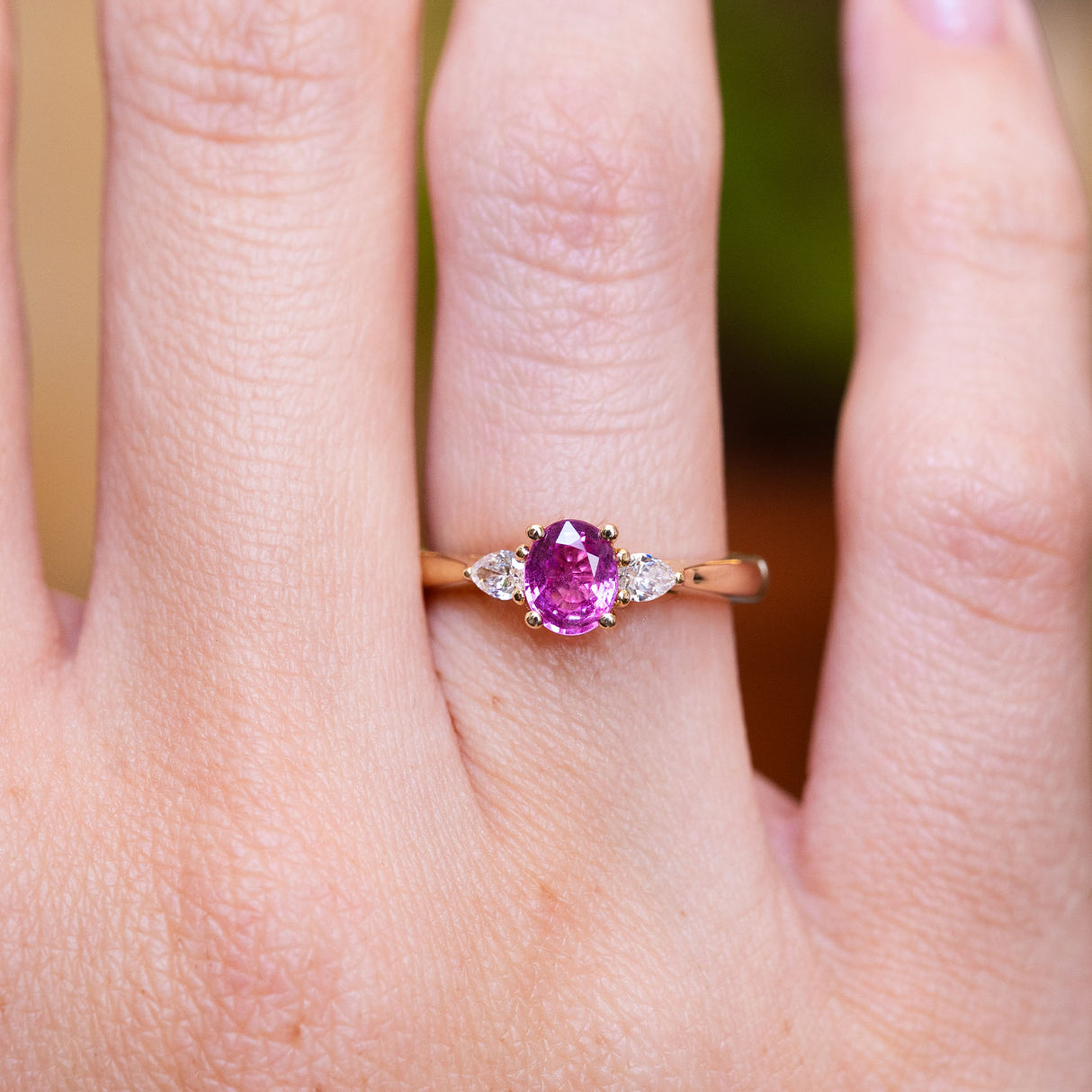 Pink oval sapphire and diamond three-stone engagement ring worn on finger with oval centre stone.