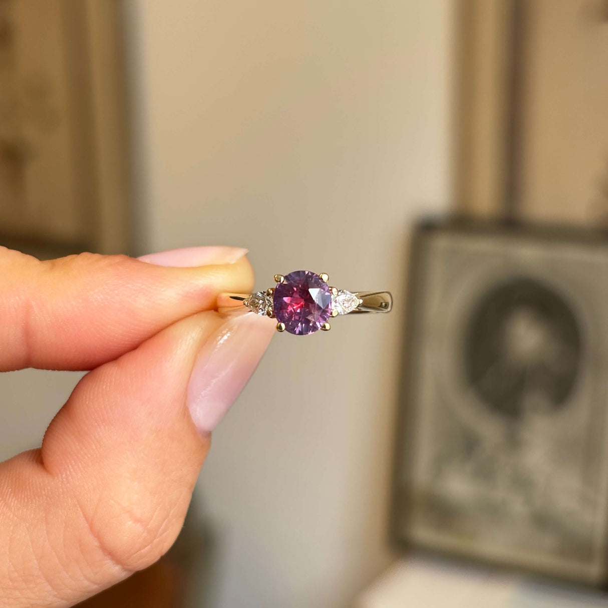Pink-red sapphire and diamond three-stone engagement ring featuring a round pink-red sapphire centre stone with pear-cut diamond side stones, held by hand.