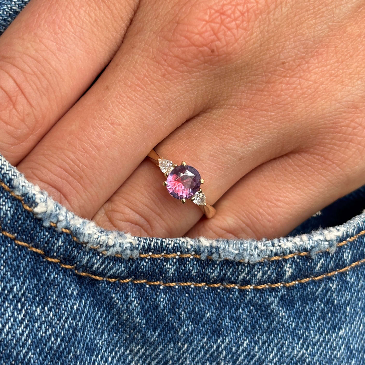 Pink-red sapphire and diamond three-stone engagement ring with a round pink-red sapphire centre stone and pear-cut diamond side stones, worn on a hand on a denim background.