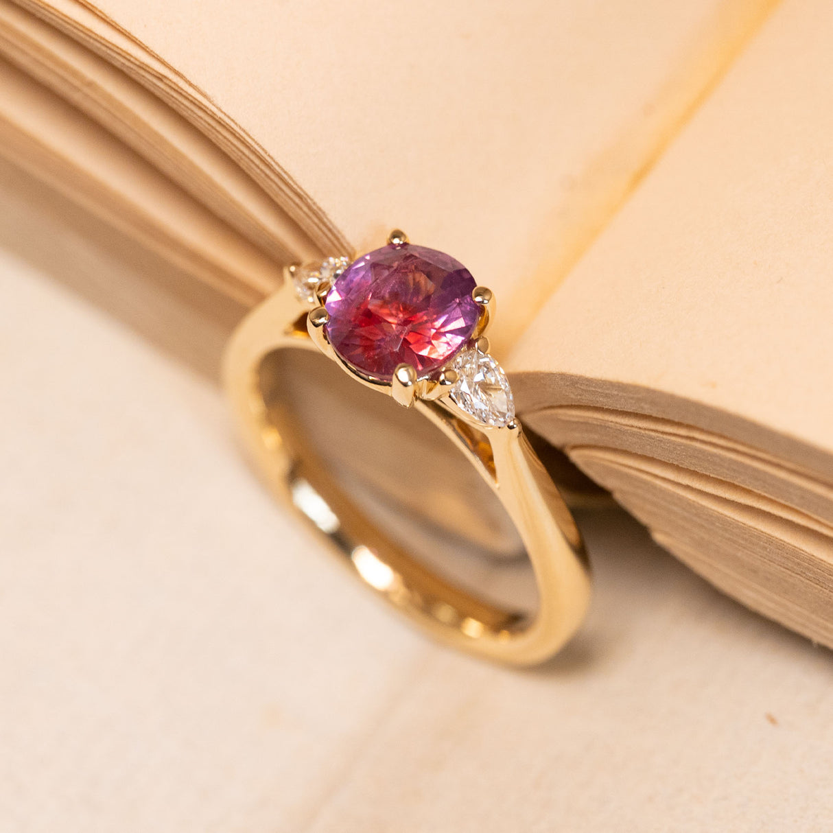 Three-stone engagement ring showcasing a richly coloured pink-red sapphire framed by pear-cut diamonds in a classic yellow gold setting.