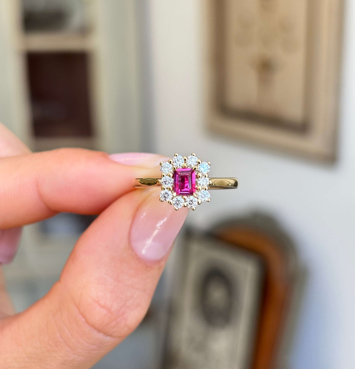 Rectangular step-cut ruby and diamond cluster engagement ring featuring a rectangular step-cut ruby surrounded by a halo of diamonds, held by hand.