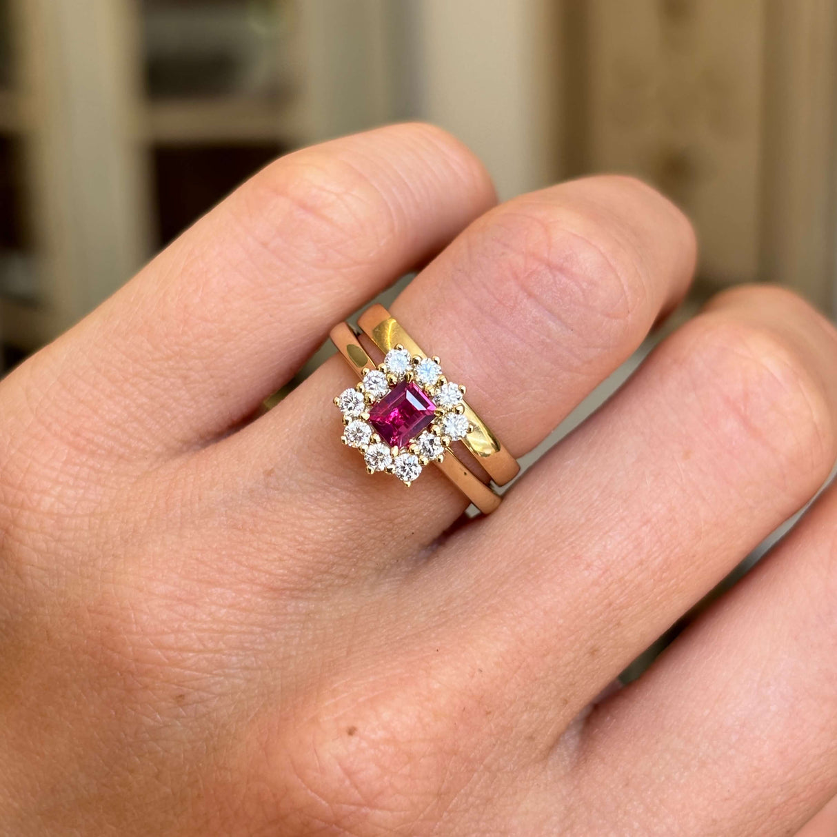 A cluster engagement ring featuring a rectangular step-cut ruby centre stone and sparkling diamond halo, worn on a finger with a gold wedding band.