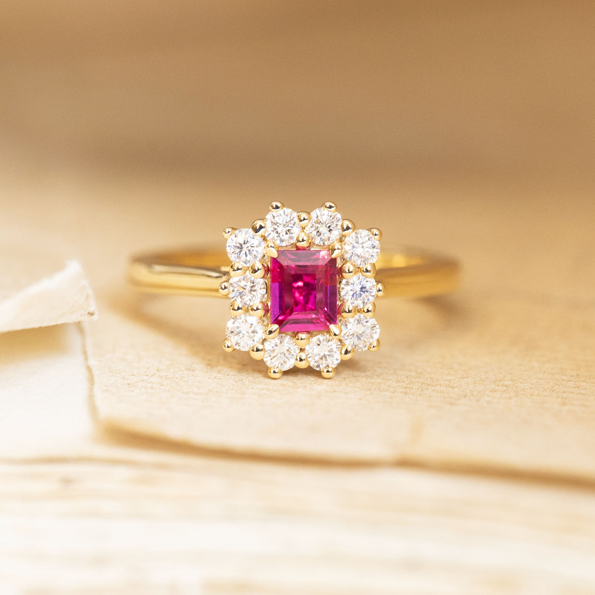 Rectangular step-cut ruby and diamond cluster engagement ring featuring a vivid red ruby framed by a sparkling diamond halo in yellow gold.