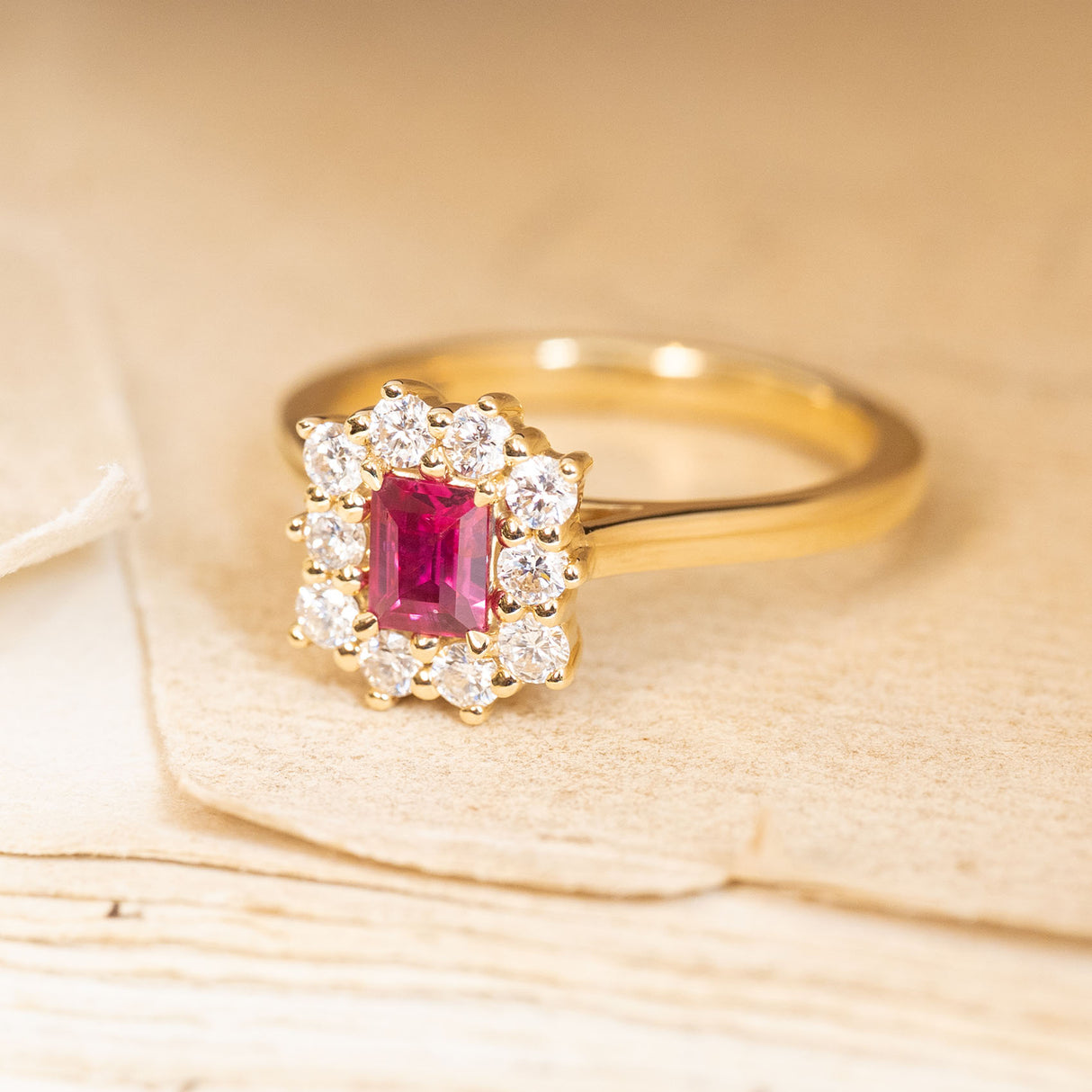 Rectangular step-cut ruby engagement ring with a diamond cluster surround and elegant yellow gold band.