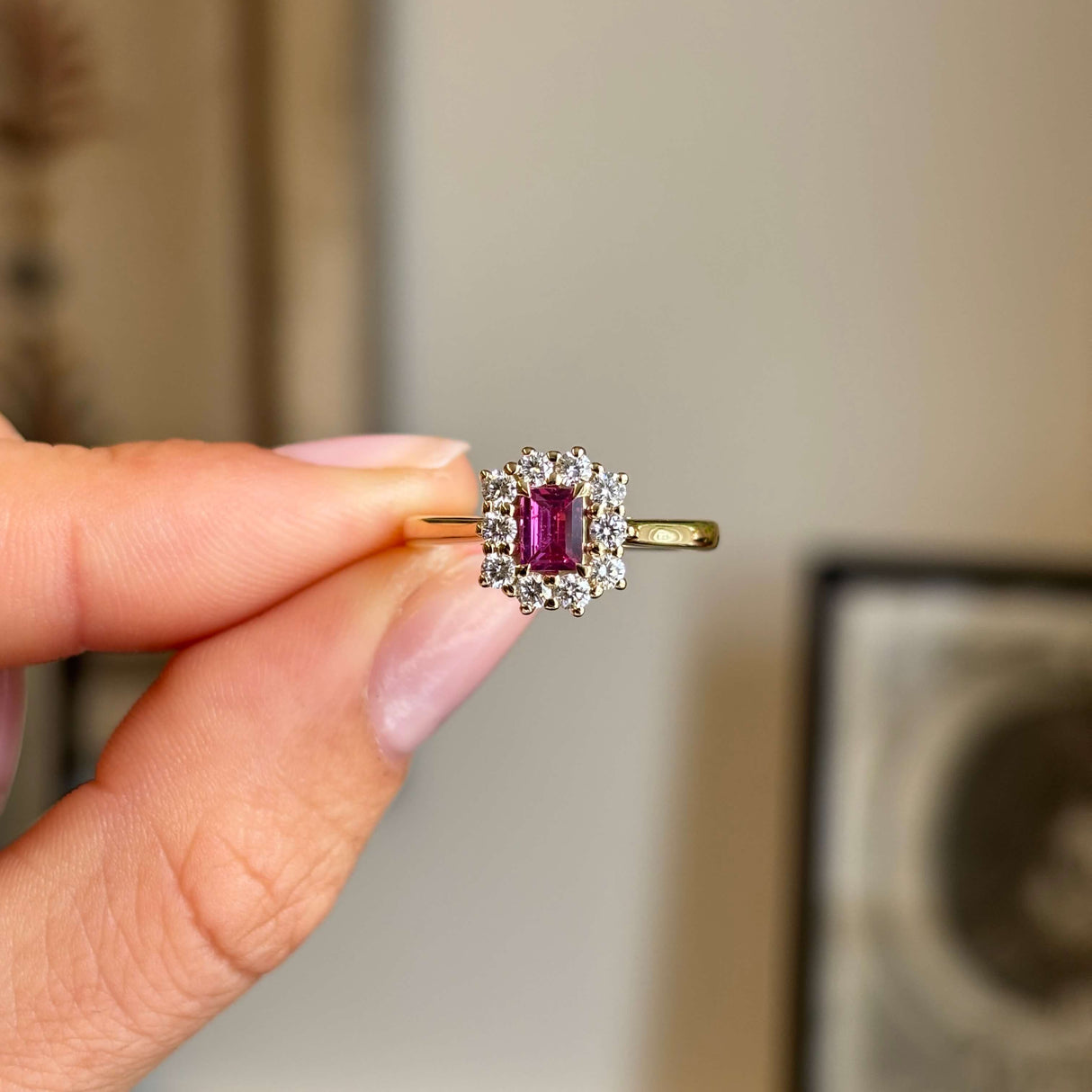 Antique-inspired step-cut pinkish-red ruby surrounded by brilliant diamonds in a yellow gold cluster setting, held between fingers against a softly blurred vintage interior background.