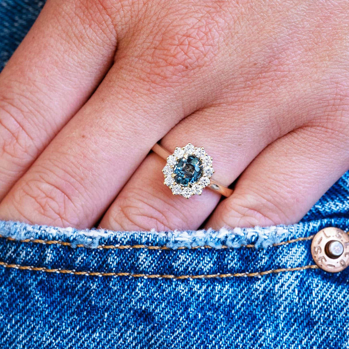 Teal sapphire cluster ring worn on hand with denim pocket detail.