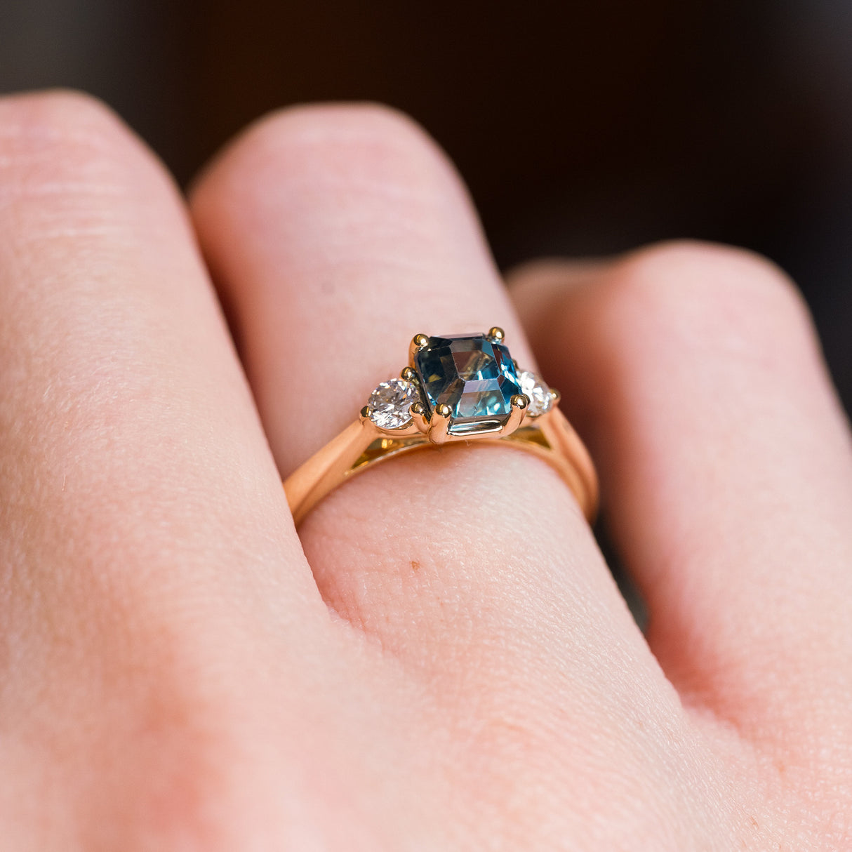 Teal sapphire and diamond three-stone engagement ring with gold band and diamond side stones.