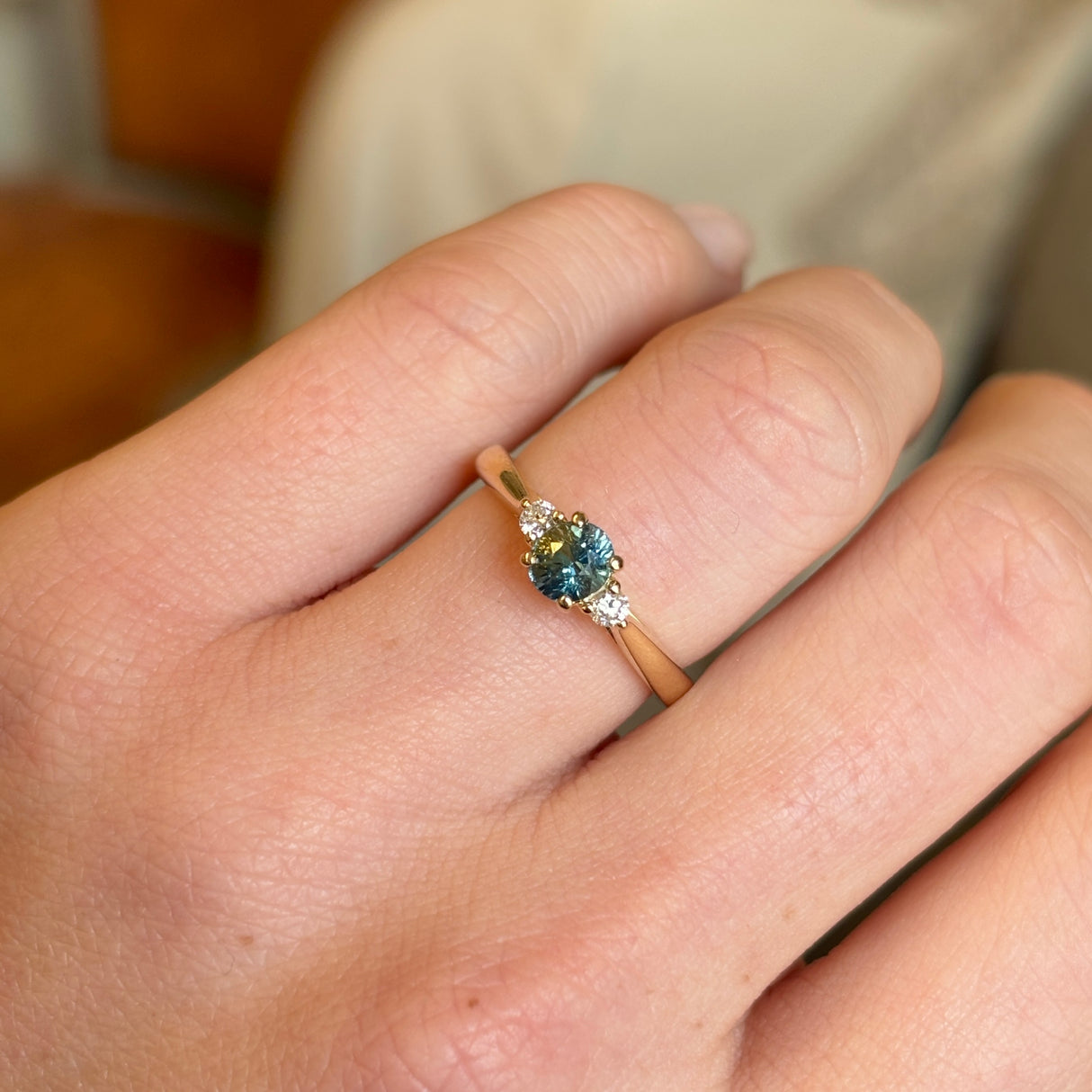 Teal sapphire and diamond three-stone engagement ring shown on hand in yellow gold setting.