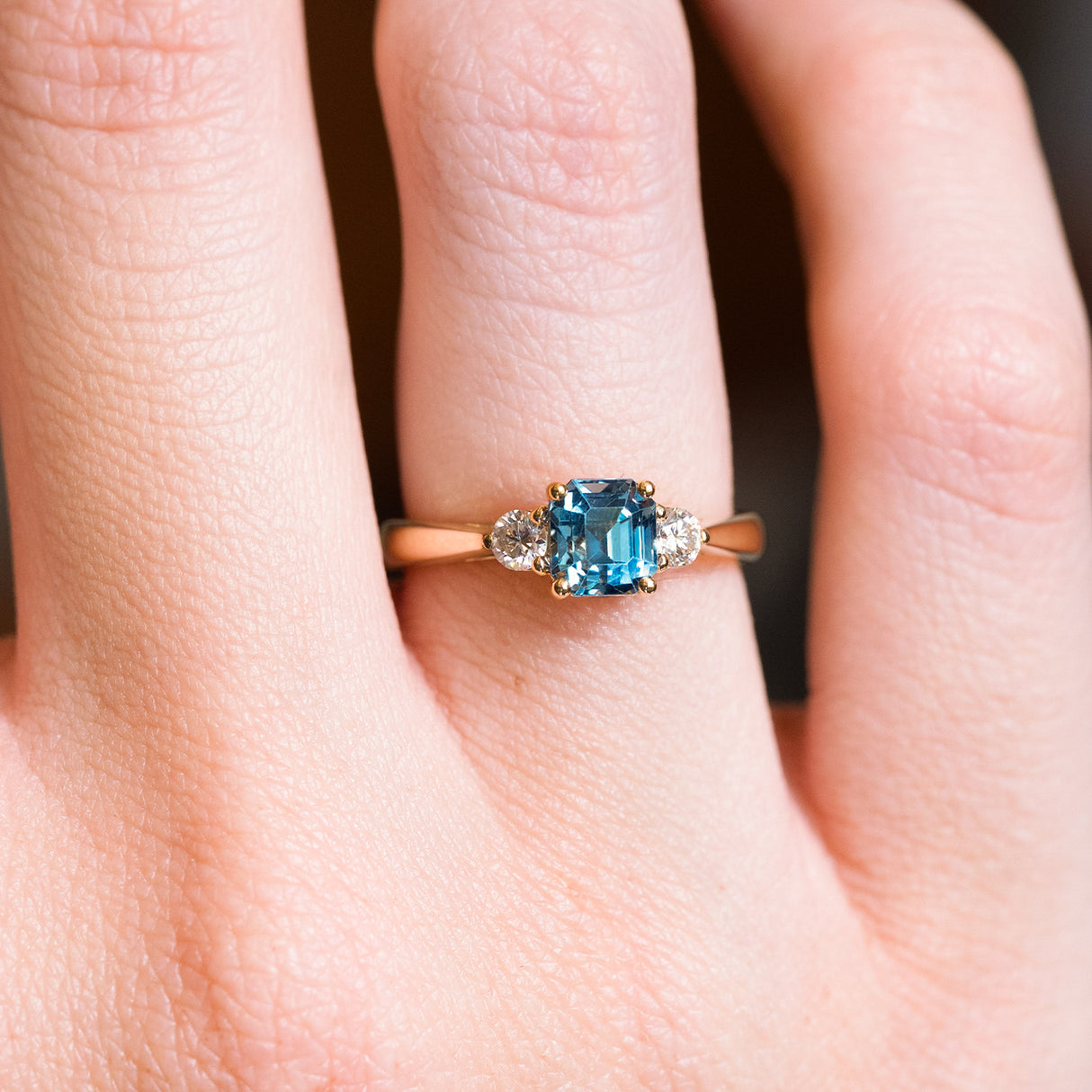 Teal sapphire and diamond three-stone engagement ring with rectangular teal sapphire centre stone worn on finger.