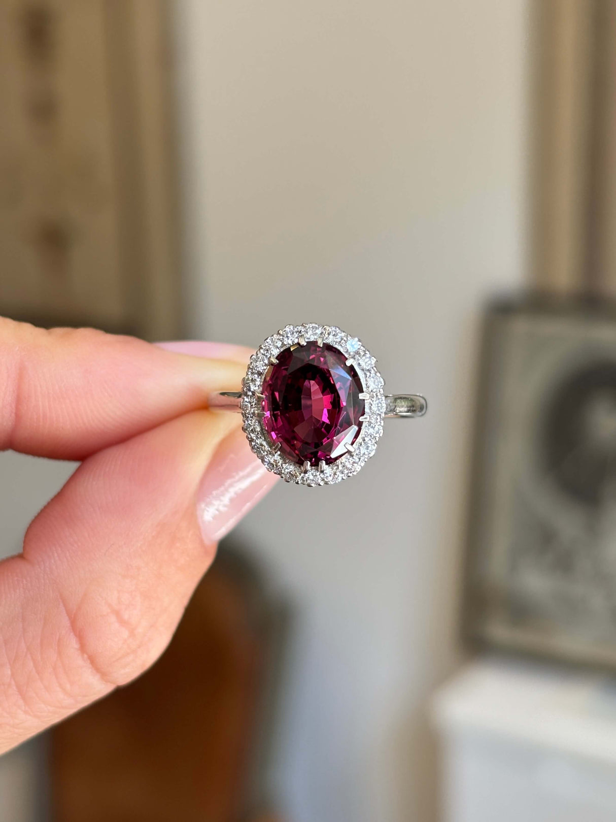 Vintage 1960s garnet and diamond cluster engagement ring featuring an oval garnet surrounded by a halo of brilliant-cut diamonds, held by hand.