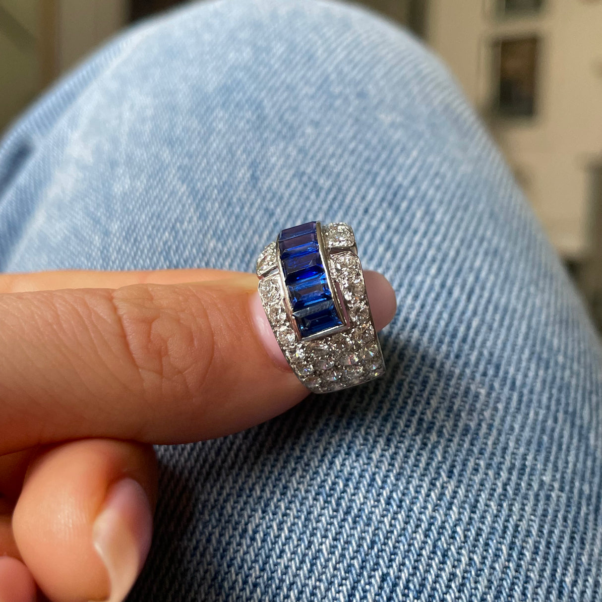 Sapphire and diamond Art Deco Band, held in fingers placed on denim jeans.