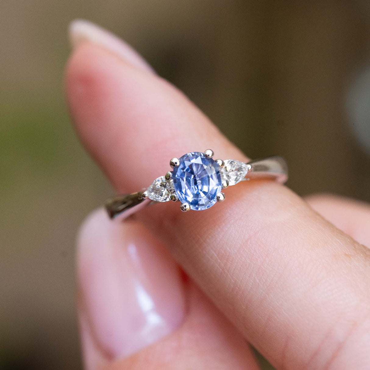 A vintage-inspired light blue sapphire and diamond three-stone engagement ring held between the fingers.