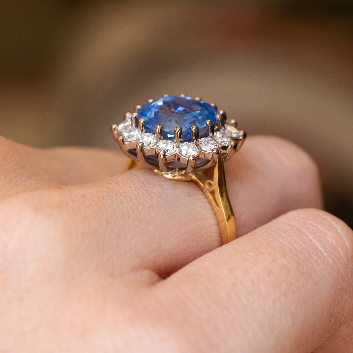 A close-up shot of a yellow and white gold ring with a large blue sapphire and a halo of diamonds, worn on a person's finger.