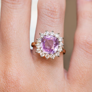 A close-up of a hand wearing an antique-inspired, pink sapphire and diamond halo cluster engagement ring with a yellow gold band.