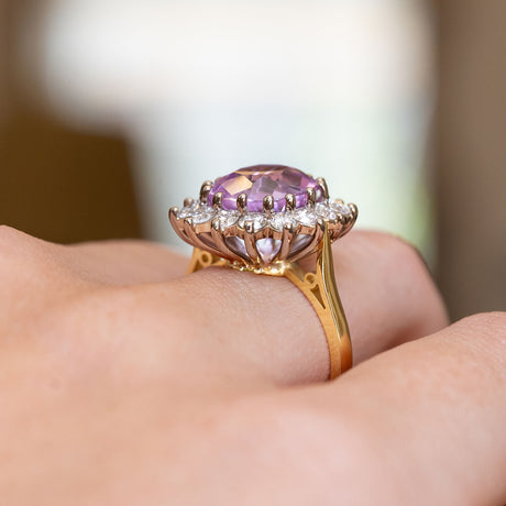 A close-up of a white and yellow gold engagement ring with a large pink sapphire at its centre surrounded by a cluster of diamonds in a claw open back setting. 