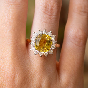 A close-up of an engagement ring worn on a finger, featuring a large cushion-cut yellow sapphire surrounded by a halo of round diamonds in a white gold setting.