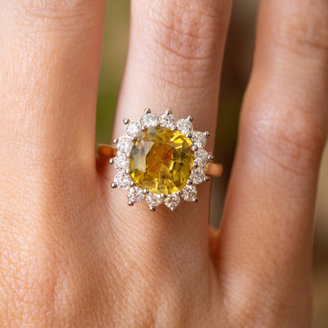 A close-up of an engagement ring worn on a finger, featuring a large cushion-cut yellow sapphire surrounded by a halo of round diamonds in a white gold setting.