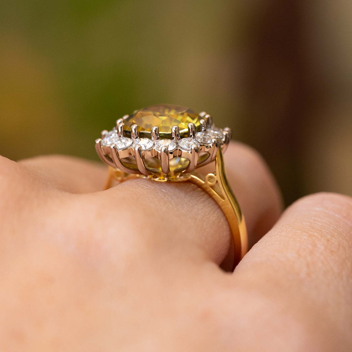 A close-up of a ring showing the yellow gold band and white gold claw-set open back setting with a bright yellow sapphire and a diamond halo.