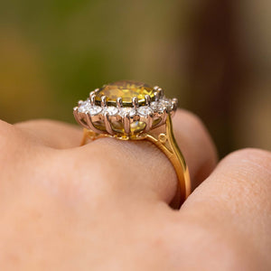 A close-up of a ring showing the yellow gold band and white gold claw-set open back setting with a bright yellow sapphire and a diamond halo.