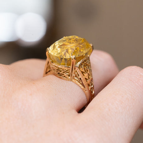 A close-up side view of a hand wearing a ring with a large yellow gemstone set in an ornate gold band.