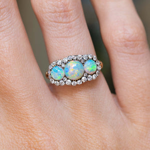 A close-up of an antique, Victorian three-stone opal & diamond cluster ring worn on hand.