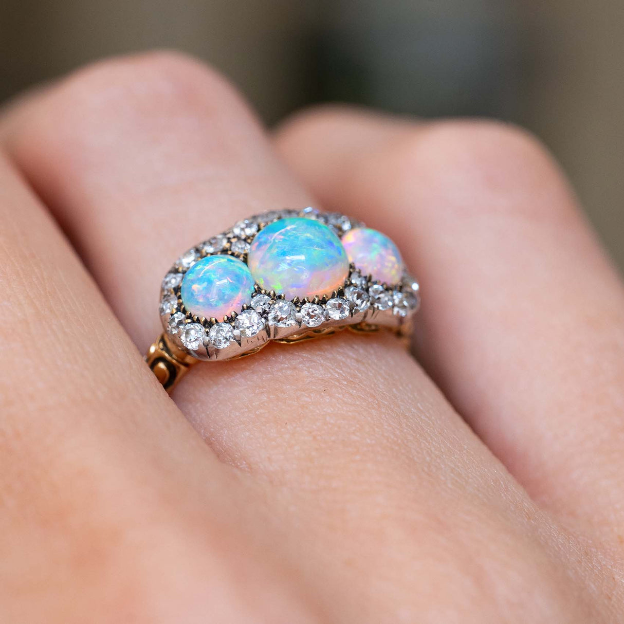 A close-up of an antique three-stone  round opal & diamond cluster ring worn on hand showing the rub-over setting and two-tone metal colour.