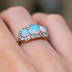 A close-up of an antique three-stone  round opal & diamond cluster ring worn on hand showing the rub-over setting and two-tone metal colour.