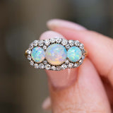 A close-up of an antique three-stone opal & diamond cluster ring shown between fingers.