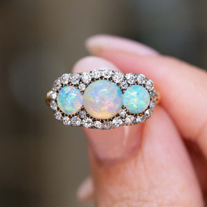 A close-up of an antique three-stone opal & diamond cluster ring shown between fingers.