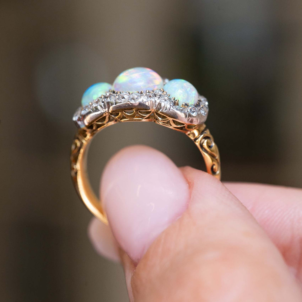 A close-up of a hand holding a gold ring with three iridescent opal gemstones and a halo of small clear stones, set against a blurred dark background.