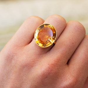 Gold ring with a large orange topaz on a hand against a blurred background.