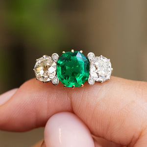 An antique Edwardian octagon-cut emerald and old-mine diamond three-stone engagement ring held between the fingers.