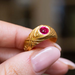 A finger holding an Antique, Art Nouveau ruby yellow gold carved solitaire ring against blurred background