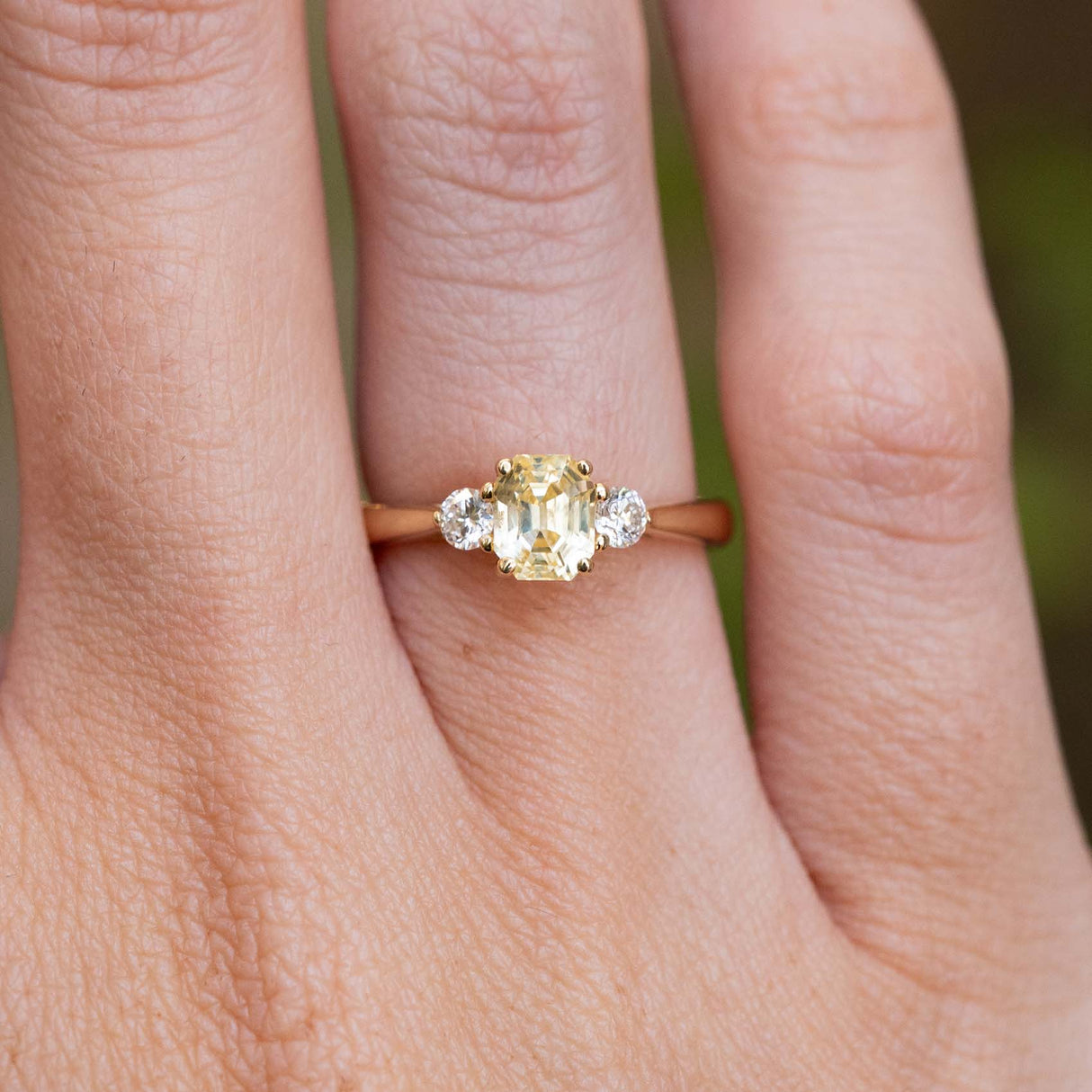 A close-up of a vintage-inspired, yellow sapphire and diamond three-stone engagement ring in yellow gold worn on a finger.