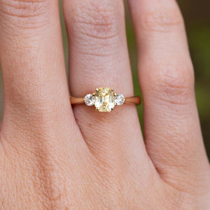 A close-up of a vintage-inspired, yellow sapphire and diamond three-stone engagement ring in yellow gold worn on a finger.