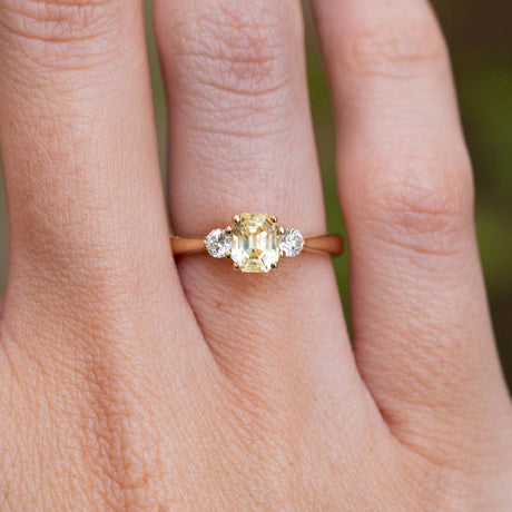 A close-up of a vintage-inspired, yellow sapphire and diamond three-stone engagement ring in yellow gold worn on a finger.
