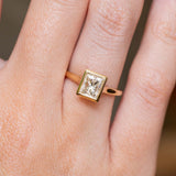 Gold Vintage-inspired, princess-cut diamond rub-over solitaire engagement ring on a person's finger against a neutral background