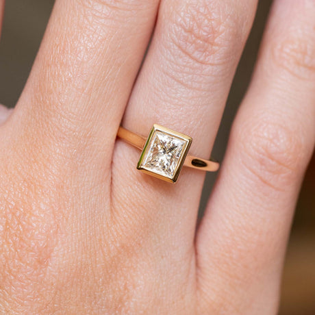 Gold Vintage-inspired, princess-cut diamond rub-over solitaire engagement ring on a person's finger against a neutral background